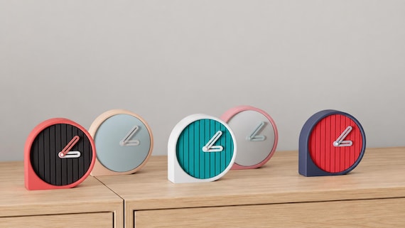 Angled close-up of Reedring Round Desk Clock examples in contrasting colors on light oak against a gray wall.