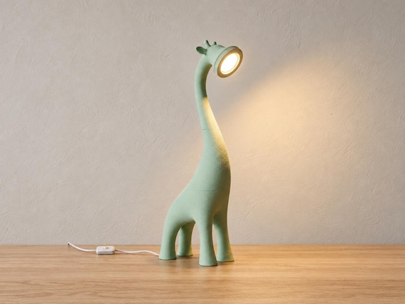 Pale green Canopy giraffe lamp glowing on a light oak desk against a neutral wall