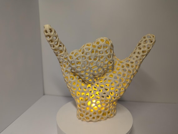 May include: A cream-colored, open-hand sculpture with a heart-shaped design in the center. The sculpture has a honeycomb-like pattern throughout, and a warm yellow light emanates from within. It is displayed on a white, circular base, creating a decorative and illuminated piece.