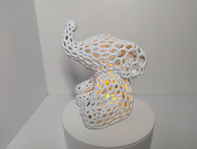 May include: A white, abstract sculpture resembling an elephant with a porous, open design. The sculpture is illuminated from within, casting a warm, yellow glow through the openings. It sits on a white, circular base, and the background is a neutral white.
