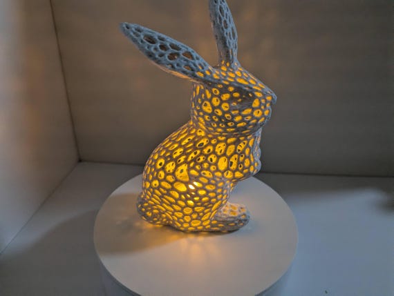 May include: A white bunny-shaped lamp with a unique, perforated design. The lamp is illuminated from within, casting a warm, yellow glow through the holes. It sits on a white, circular base, creating a soft, ambient light. The bunny's ears are upright, and its body is in a sitting position.