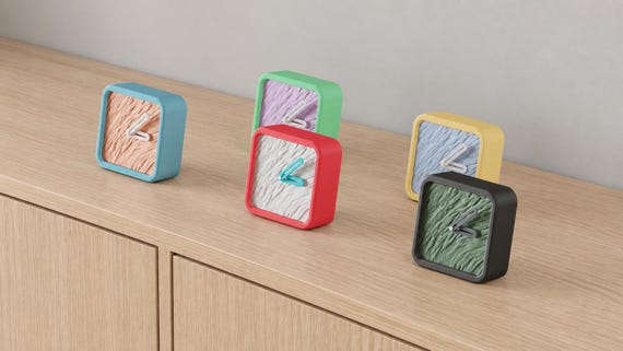 Angled close-up of Meadowgrain Textured Desk Clock examples in contrasting colors on light oak against a gray wall.