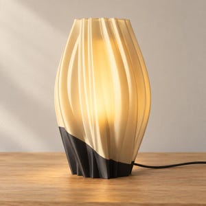 May include: A table lamp with a translucent, cream-colored, vertically ribbed shade that emits a warm glow. The base of the lamp is solid black and features a matching vertical, rippled texture that seamlessly continues the pattern of the shade.