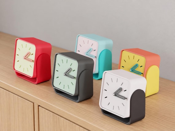Angled close-up of Pivotline Desk Clock examples in contrasting colors on light oak against a gray wall.