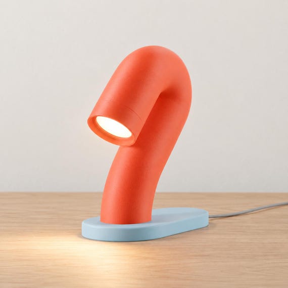 Citrus Tide Swayarc lamp with an orange curved body, pale blue oval base and warm LED light on a wood desk, with its power cord visible.
