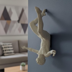 Könnte beinhalten: Eine cremefarbene Wandskulptur, die eine Figur in einer Tauchpose darstellt. Die Skulptur ist an einer blauen Wand befestigt. Die Beine der Figur sind erhoben und die Arme ausgestreckt. Die Skulptur hat eine strukturierte Oberfläche.