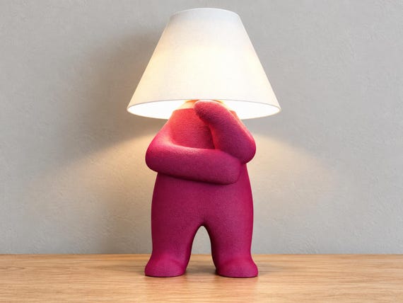 Magenta character lamp in a thinking pose with white smooth shade, close-up three-quarter view on light oak against a pale gray wall.