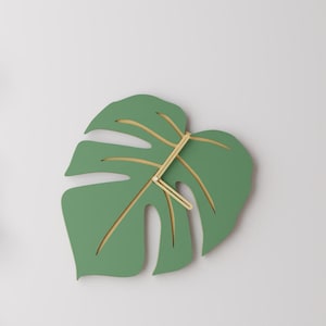 May include: A wall clock shaped like a green monstera leaf with cutouts revealing a light wood-toned base. The clock features two thin, light-colored wooden hands positioned at the center, extending outward to indicate the time on the leaf surface.