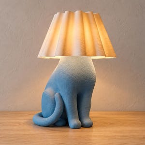 Pode incluir: Um candeeiro de mesa em forma de gato sentado com um acabamento azul claro texturizado. O candeeiro apresenta um abajur cónico plissado em branco sujo que emite um brilho quente e suave, iluminando a base de cima para baixo.