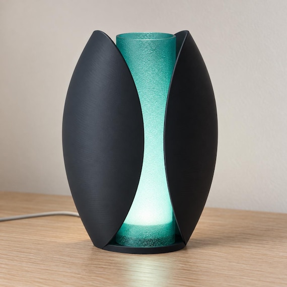 Midnight Tide Lunafold lamp with Charcoal curved outer panels and a glowing Teal inner shade on a pale wood tabletop, viewed from the front.
