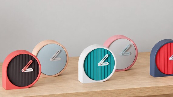 Alternate close-up of Reedring Round Desk Clock examples on light oak, showing their colored faces, hands and body profiles.
