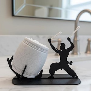 Può includere: Un porta-cotton fioc originale con la silhouette nera di un artista marziale che colpisce un tamburo. Il supporto a forma di tamburo è grigio chiaro con una superficie strutturata ed è sostenuto da un supporto nero. Due cotton fioc sono tenuti dall'artista marziale.