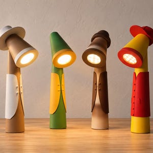 May include: Four cylindrical LED desk lamps shaped like figures wearing hats and vests. The first is tan with a white vest and hat, the second is green with a yellow vest, the third is brown with a dark brown vest, and the fourth is yellow with a red vest and cap.
