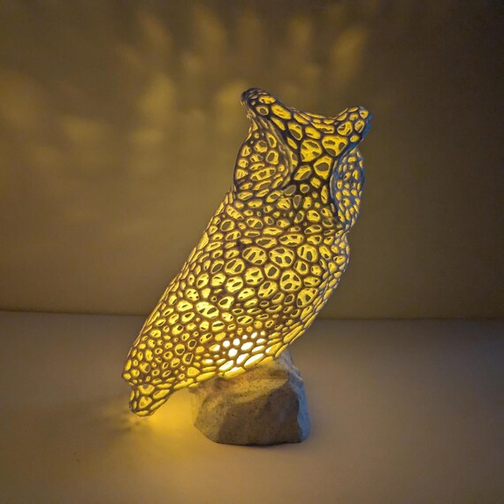 May include: A decorative owl lamp with a unique design. The owl is primarily yellow with a pattern of dark spots, and it sits atop a gray stone base. The lamp emits a warm, inviting glow, highlighting the intricate details of the owl's form.