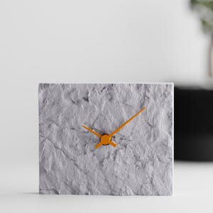 May include: A rectangular clock with a textured, light gray stone-like face and bright orange hour and minute hands. The clock features a rugged, uneven surface that mimics natural rock, providing a stark contrast to the smooth, vibrant orange hands.