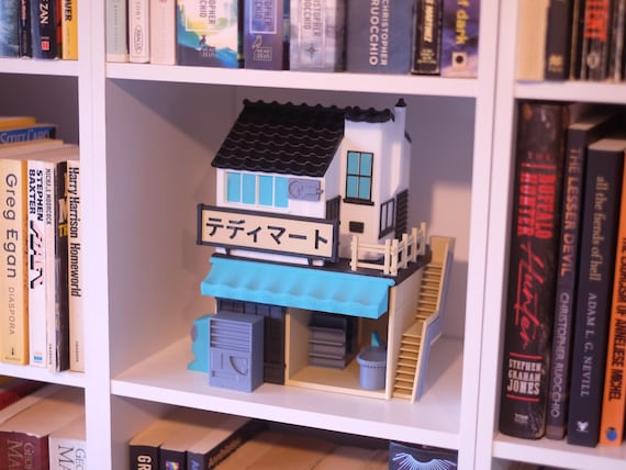 Multicolor miniature neighborhood shop with dark tiled roof, blue awning and outdoor stairs on a bookshelf.