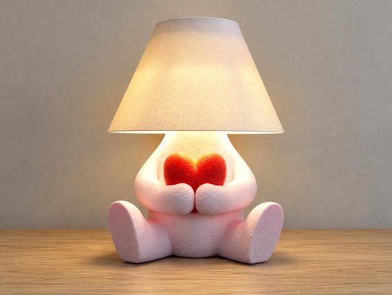 Pink character lamp holding a red heart beneath a white smooth shade, close-up three-quarter view on light oak against a pale gray wall.