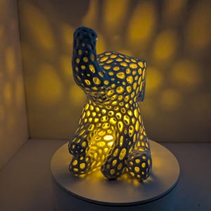 May include: A blue and white ceramic sculpture of an abstract animal form, illuminated from within. The sculpture has a porous design, allowing light to shine through. It sits on a white circular base, with a warm yellow glow emanating from the interior.