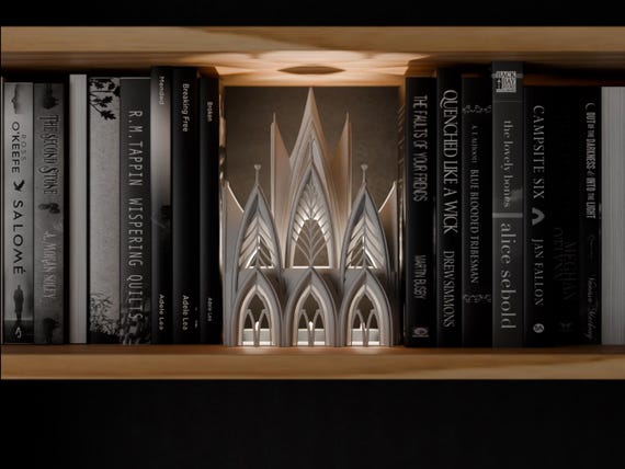 Lacelight Cathedral Book Nook in a light neutral color, shown from the front between books on a wooden shelf.