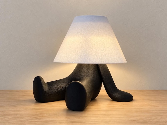 Black reclining character lamp with extended legs and white smooth shade, close-up three-quarter view on light oak against a pale gray wall.
