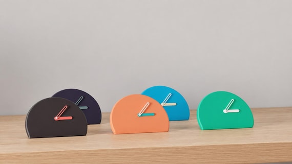 Alternate close-up of Halfmoon Folded Desk Clock examples on light oak, showing their colored faces, hands and body profiles.