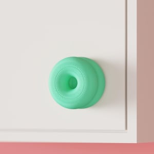 Puede incluir: Un pomo de armario de color verde menta con forma de dónut, acabado mate liso y una abertura circular central, montado sobre una superficie rectangular blanca y plana. El pomo presenta un perfil redondeado y escalonado hacia el centro.