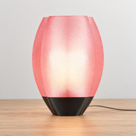 Veilcrest lamp with a glowing Cherry Pink translucent shade and black base, viewed from the front on a pale wood tabletop with its cord visible.