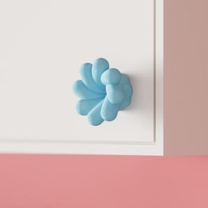 May include: A light blue cabinet knob with a rounded, multi-lobed flower-like shape. The knob is attached to a flat, white cabinet door surface. The design features smooth, curved segments that radiate from the center point of the fixture.