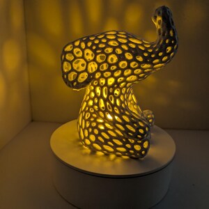 May include: A unique, abstract lamp with a porous, textured design. The lamp is dark gray with numerous circular openings that emit a warm, yellow light. It sits on a white circular base, creating a striking contrast.