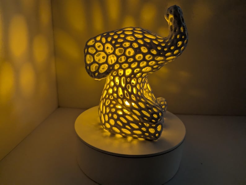 May include: A unique, abstract lamp with a porous, textured design. The lamp is dark gray with numerous circular openings that emit a warm, yellow light. It sits on a white circular base, creating a striking contrast.