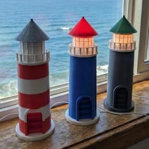 May include: Three decorative lighthouse lamps with red, blue, and gray bodies. The red and white striped lighthouse has a gray roof. The blue lighthouse has a red roof, and the gray lighthouse has a green roof. Each lamp has a white base and a small door.