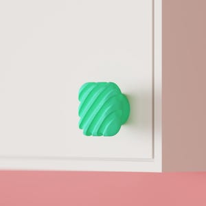 May include: A square cabinet knob in a bright mint green color featuring a series of diagonal, raised ridges across its front surface. The knob is mounted onto a flat, white panel, showcasing a textured, geometric design with rounded corners.