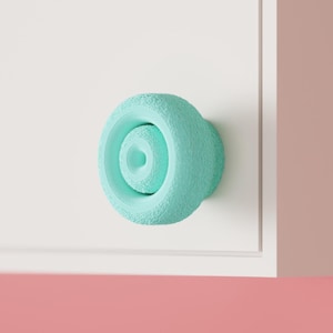May include: A round, mint green cabinet knob with a textured, granular outer surface and a smooth, recessed center. The knob is mounted on a flat, white surface, featuring a circular design that protrudes from the front of the cabinet door.