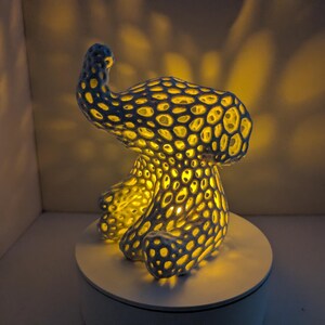 May include: A unique, blue and yellow decorative lamp with a porous, organic design. The lamp is illuminated from within, casting a warm yellow glow through the intricate pattern. It sits on a white circular base.