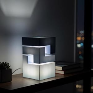 May include: A contemporary geometric table lamp with a layered design. The lamp has a black top, a grey middle section, and a white base that emits a soft glow. The lamp is on a wooden surface, with a small potted plant and books nearby.