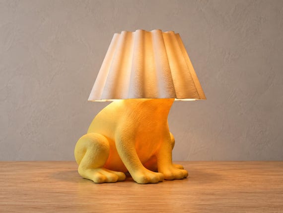 Golden yellow crouching frog lamp with white curved fluted shade, close-up three-quarter view on light oak against a pale gray wall.