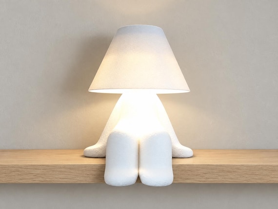 White character lamp with dangling feet and white smooth shade, close-up on an oak shelf against a pale gray wall.