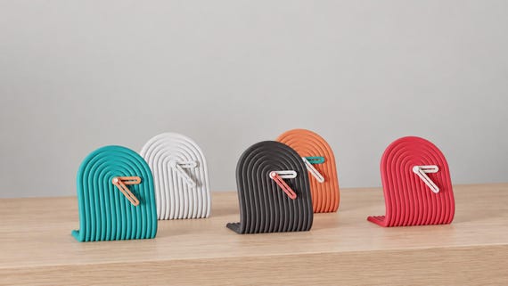 Alternate close-up of Archwave Sculptural Desk Clock examples on light oak, showing their colored faces, hands and body profiles.