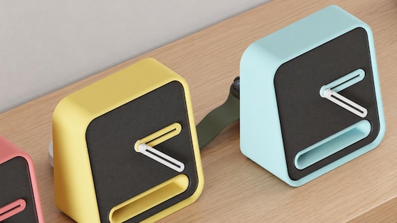 Angled close-up of Slothour Desk Clock examples in contrasting colors on light oak against a gray wall.