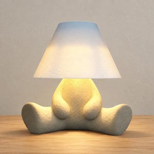 May include: A table lamp featuring a textured, off-white base shaped like a seated figure with outstretched legs. The lamp is topped with a conical, white lampshade that emits a warm, soft glow, illuminating the textured surface of the base.
