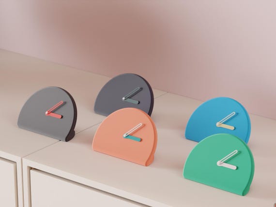 Halfmoon Folded Desk Clock examples in contrasting colors on a pale cabinet, showing the finished clock faces and hands.
