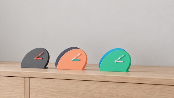 Angled close-up of Halfmoon Folded Desk Clock examples in contrasting colors on light oak against a gray wall.