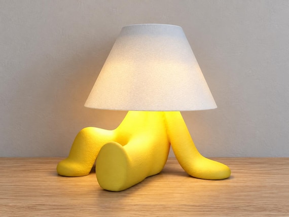 Yellow reclining character lamp with one bent knee and white smooth shade, close-up three-quarter view on light oak against a pale gray wall.