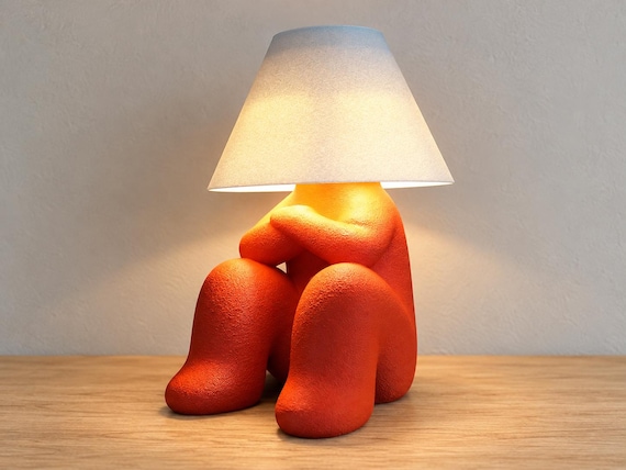 Orange character lamp with folded arms and raised knees beneath a white smooth shade, close-up three-quarter view on light oak against a pale gray wall.