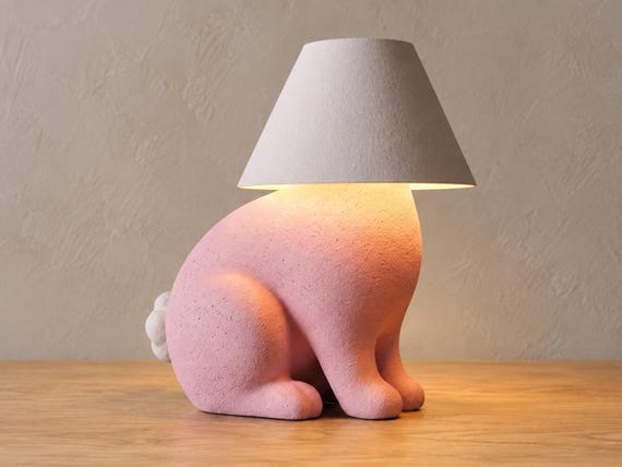 Pink rabbit lamp with white tail and smooth white shade, close-up three-quarter view on light oak against a pale gray wall.