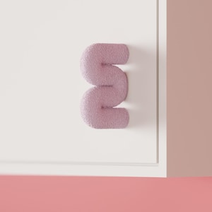 Peut inclure: Une poignée de placard en forme de S, de couleur rose poudré et à la texture granuleuse, fixée sur une porte de placard blanche et plate. La poignée présente une forme tubulaire arrondie qui contraste avec la finition lisse et mate de la porte.