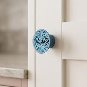 May include: A circular cabinet knob in a light blue color featuring a raised, intricate, brain-like coral pattern on its front face. The knob has a cylindrical base and is mounted onto a flat, off-white vertical panel surface.