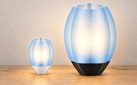 Two Ice Blue Veilcrest lamps on a wood tabletop: smaller lamp with white base beside a lamp twice as tall with black base, both illuminated.