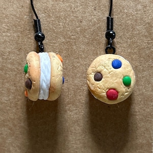 May include: A pair of novelty earrings featuring a cookie theme. One earring is a cookie sandwich with white filling and colorful candy pieces. The other earring is a round cookie with colorful candy pieces. Both earrings have black ear wires.