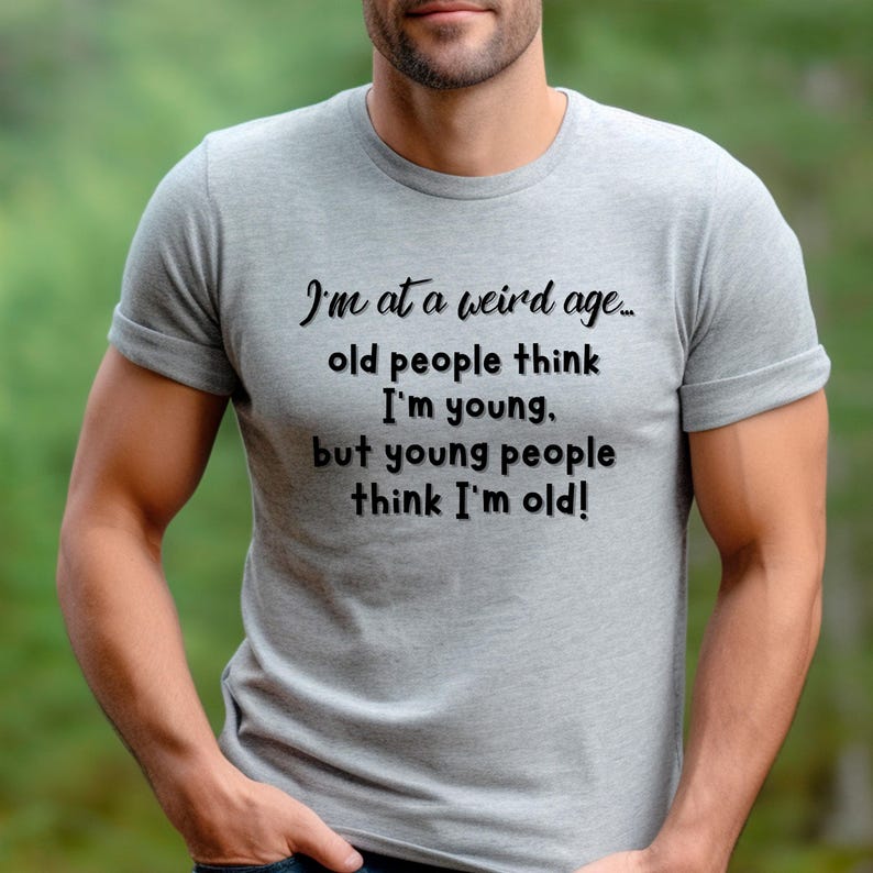 Puede incluir: Una camiseta gris con texto negro que dice "Estoy en una edad extra&ntilde;a... la gente mayor piensa que soy joven. pero la gente joven piensa que soy viejo!"